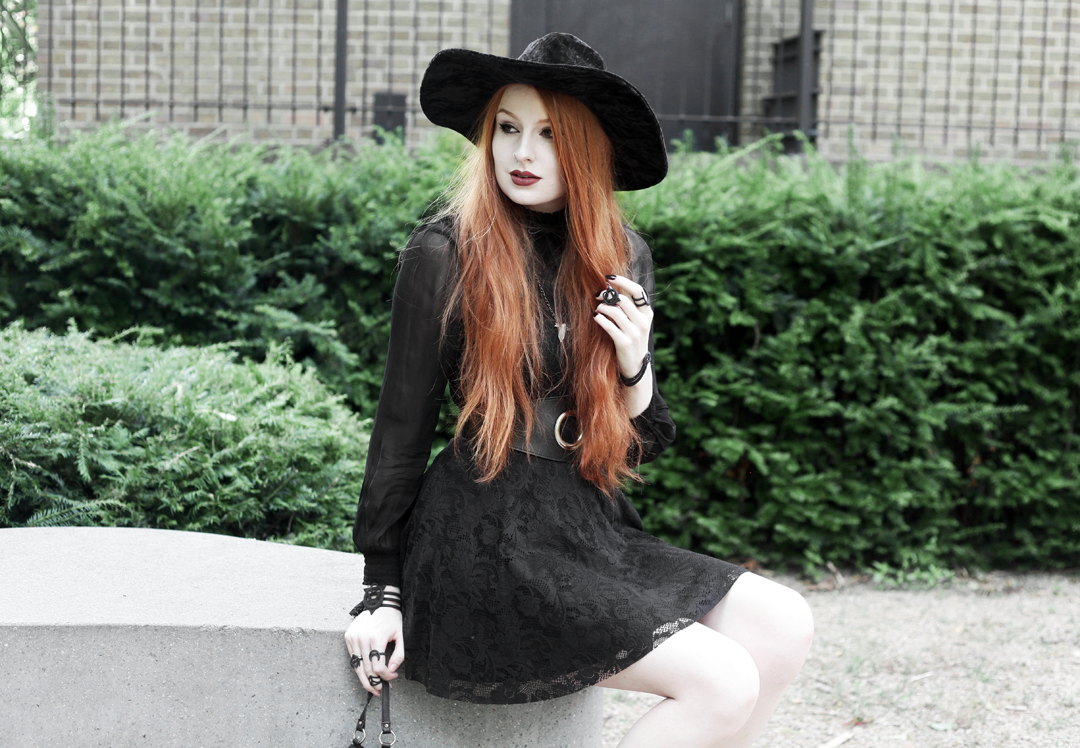 Olivia Emily wears Asos Sheer Shirt, Black Milk Black Lace Skater Dress, Killstar Velvet Witch Brim Hat, Zana Bayne Cincher Waist Belt, Rogue and Wolf Matte Black Bracelet & Rings, and Asos Ariana Lace Up Boots